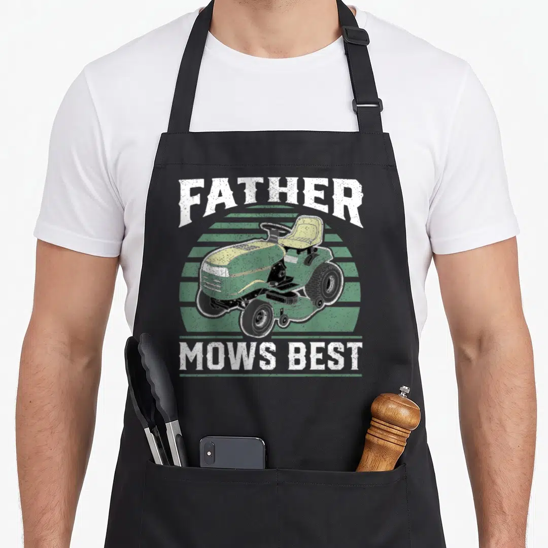 Father mower apron - Retro riding lawn mower illustration with FATHER MOWS BEST text on green stripes — shown on a black cotton canvas apron against a white background