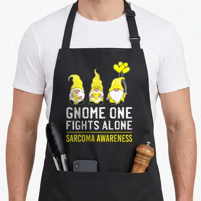 Fight alone ribbon apron - Three yellow gnomes holding balloons above white and yellow text reading GNOME ONE FIGHTS — shown on a black cotton canvas apron against a white background