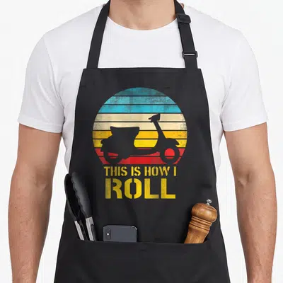 Moped scooter apron - Grey scooter silhouette on yellow blue red striped circle with THIS IS HOW I ROLL text — shown on a black cotton canvas apron against a white background