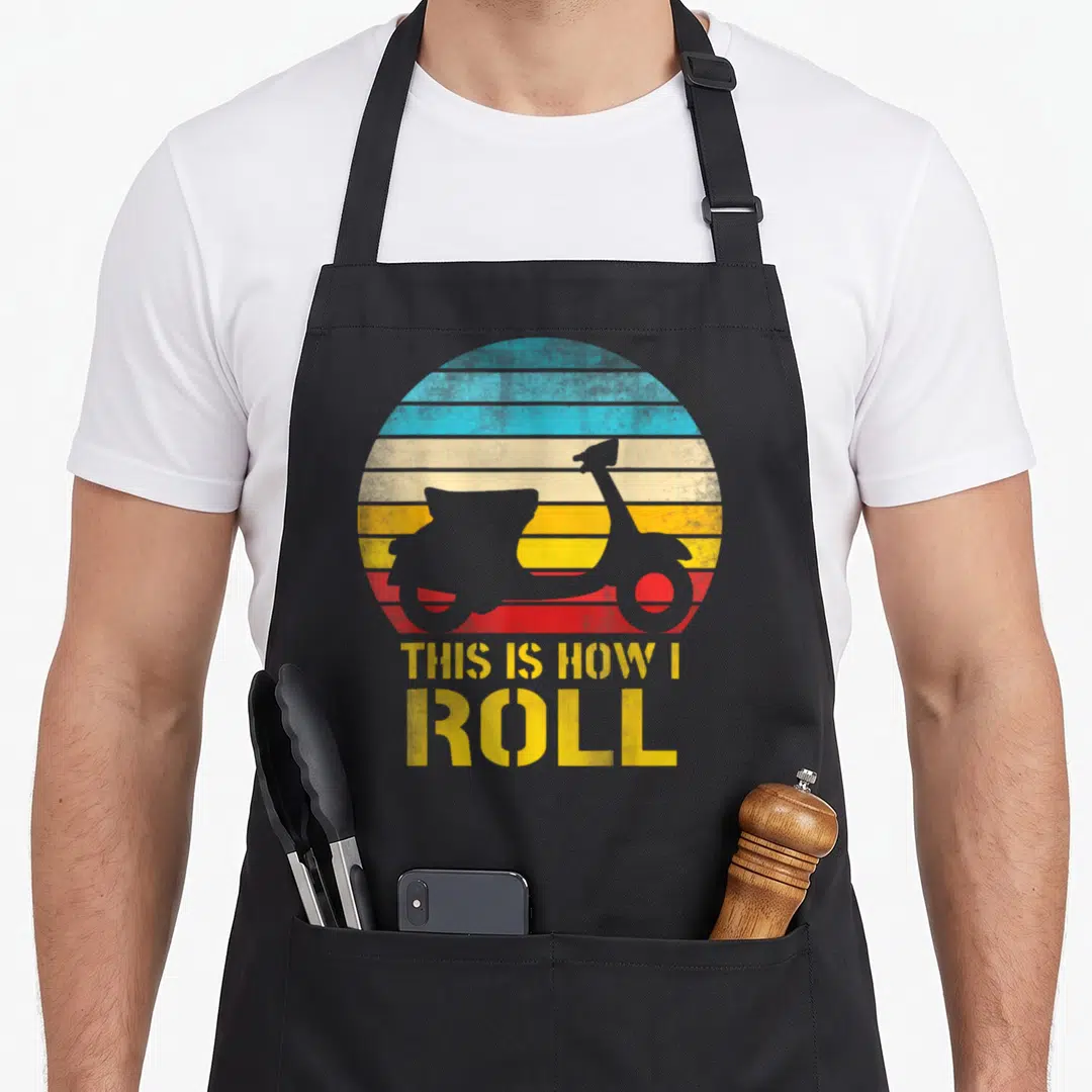 Moped scooter apron - Grey scooter silhouette on yellow blue red striped circle with THIS IS HOW I ROLL text — shown on a black cotton canvas apron against a white background
