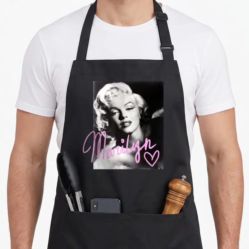 Marilyn novelty apron - Black white Marilyn Monroe photo with pink script name and heart overlay — shown on a black cotton canvas apron against a white background