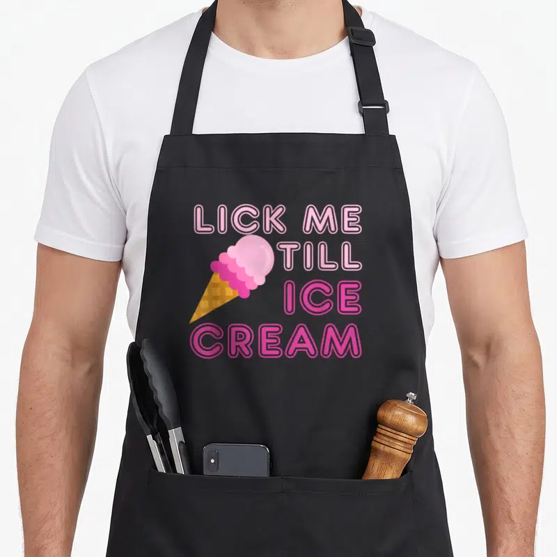 Lick ice cream apron - Pink cartoon ice cream cone with LICK ME TILL ICE CREAM text in pink and orange outlines — shown on a black cotton canvas apron against a white background