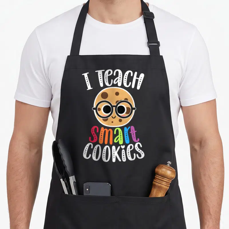 Teach cookies apron - Cartoon cookie with glasses and text I Teach Smart Cookies in white and multi-colored letters — shown on a black cotton canvas apron against a white background