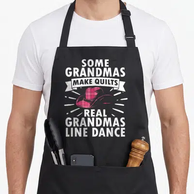 Grandma teacher apron - Pink plaid cowboy hat with white text reading Some Grandmas Make Quilts Real Grandmas Line Dance — shown on a black cotton canvas apron against a white background