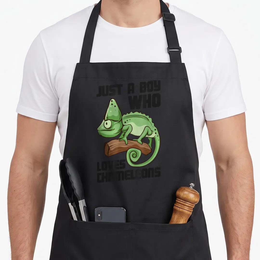 Funny lizard chameleon apron - Cartoon green chameleon on brown log with text JUST A BOY WHO LOVES CHAMELEONS — shown on a black cotton canvas apron against a white background
