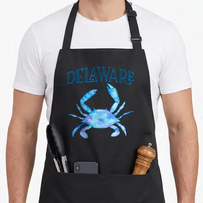Delaware crab apron - Blue watercolor crab centered below arched DELAWARE text in blue — shown on a black cotton canvas apron against a white background