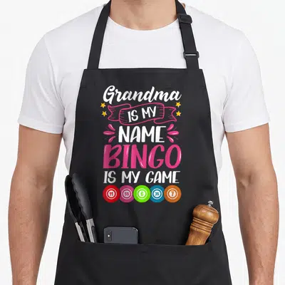 Grandma bingo apron - Stacked text Grandma IS MY NAME BINGO IS MY GAME with five colored numbered balls — shown on a black cotton canvas apron against a white background