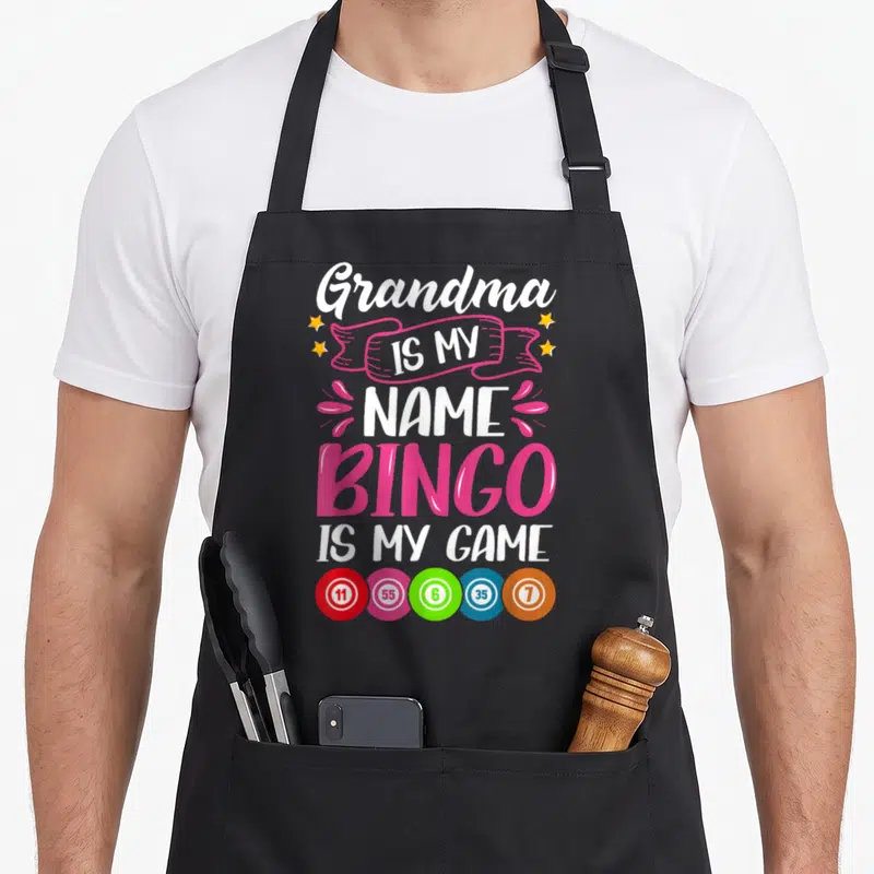 Grandma bingo apron - Stacked text Grandma IS MY NAME BINGO IS MY GAME with five colored numbered balls — shown on a black cotton canvas apron against a white background