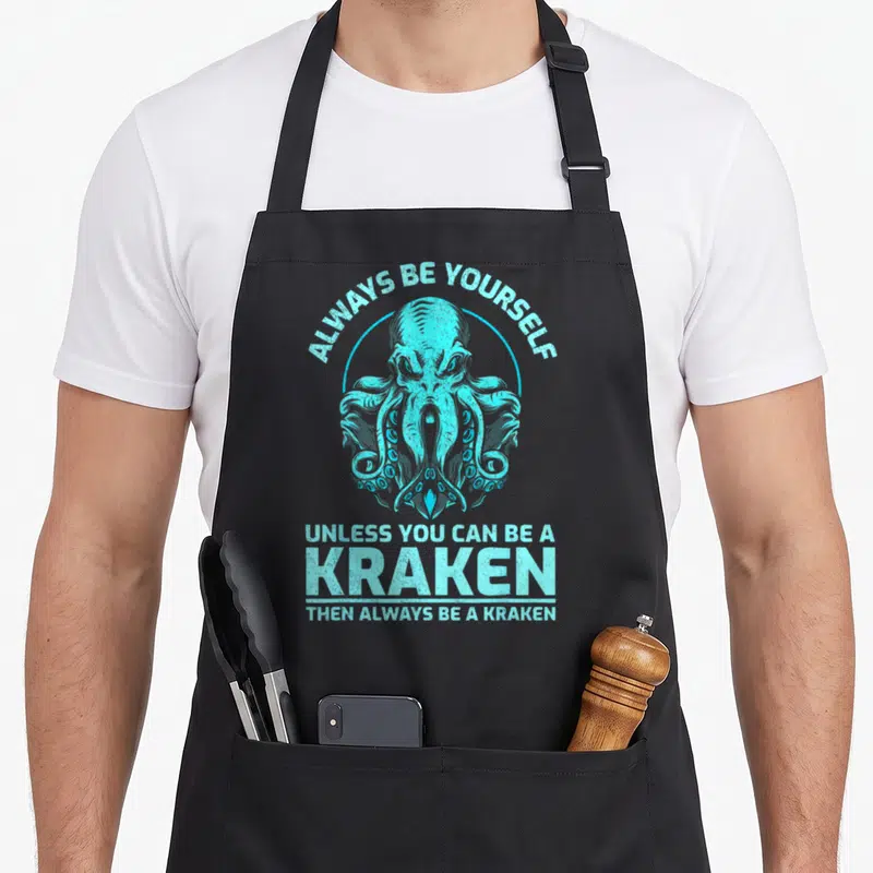 Yourself kraken apron - Cyan kraken head in circle with arched text above and stacked text below — shown on a black cotton canvas apron against a white background