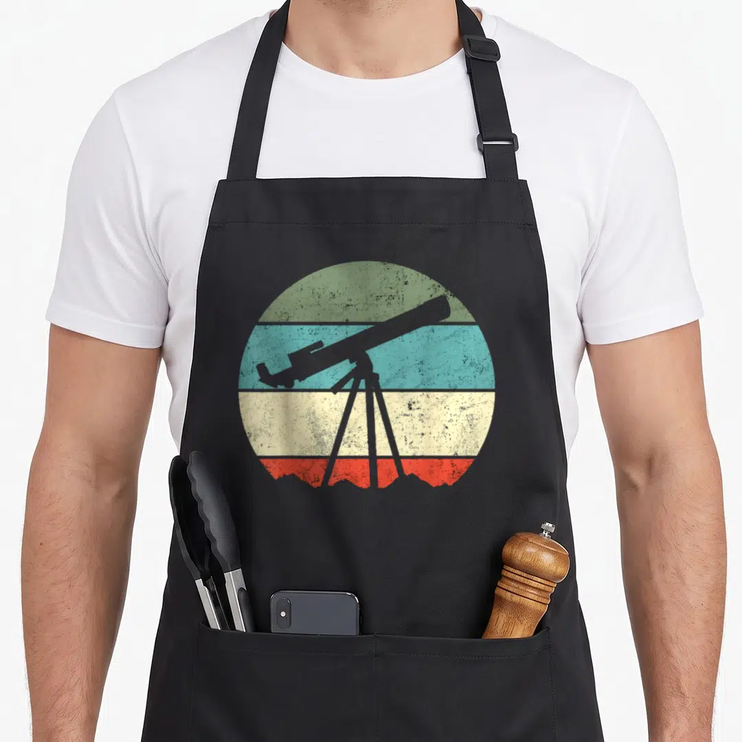 Astronomer telescope lover apron - Grey telescope silhouette over green blue beige and red horizontal bands in a circle — shown on a black cotton canvas apron against a white background