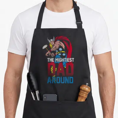 Mightiest father day apron - Thor cartoon figure above stacked text reading The Mightiest Dad Around in red, blue — shown on a black cotton canvas apron against a white background