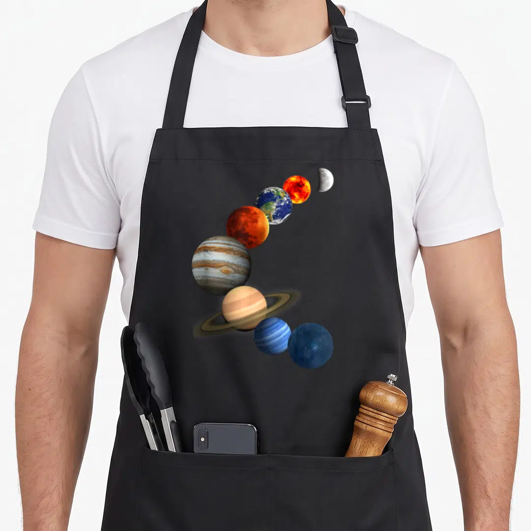 Our space planets apron - Realistic planets and moon in curved diagonal line using blue orange brown grey — shown on a black cotton canvas apron against a white background