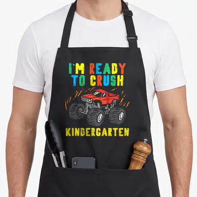 Kindergarten monster truck apron - Red monster truck with orange splatter and multicolored text reading ready to crush — shown on a black cotton canvas apron against a white background