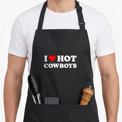 Hot western aprons - White text I HOT COWBOYS with a red heart symbol centered on the chest panel — shown on a black cotton canvas apron against a white background