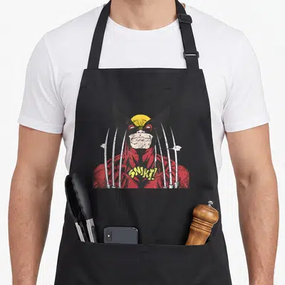 Snikt hobby apron - Wolverine cartoon in red yellow black white with SNIKT text and six claws — shown on a black cotton canvas apron against a white background