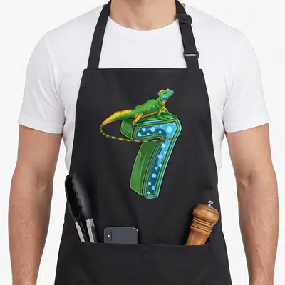 Reptile birthday apron - Cartoon iguana in green blue and yellow draped over a 3D number 7 — shown on a black cotton canvas apron against a white background