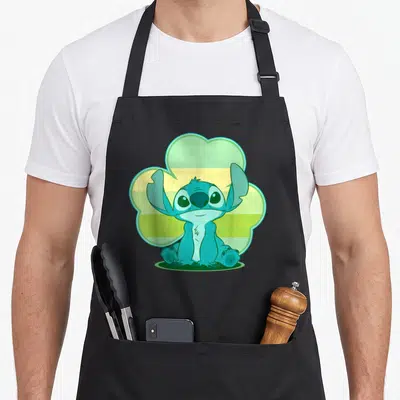 Stitch st patrick day aprons - Blue cartoon creature sitting in front of a green and yellow striped cloud shape — shown on a black cotton canvas apron against a white background