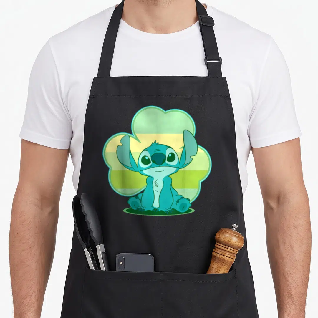 Stitch st patrick day aprons - Blue cartoon creature sitting in front of a green and yellow striped cloud shape — shown on a black cotton canvas apron against a white background