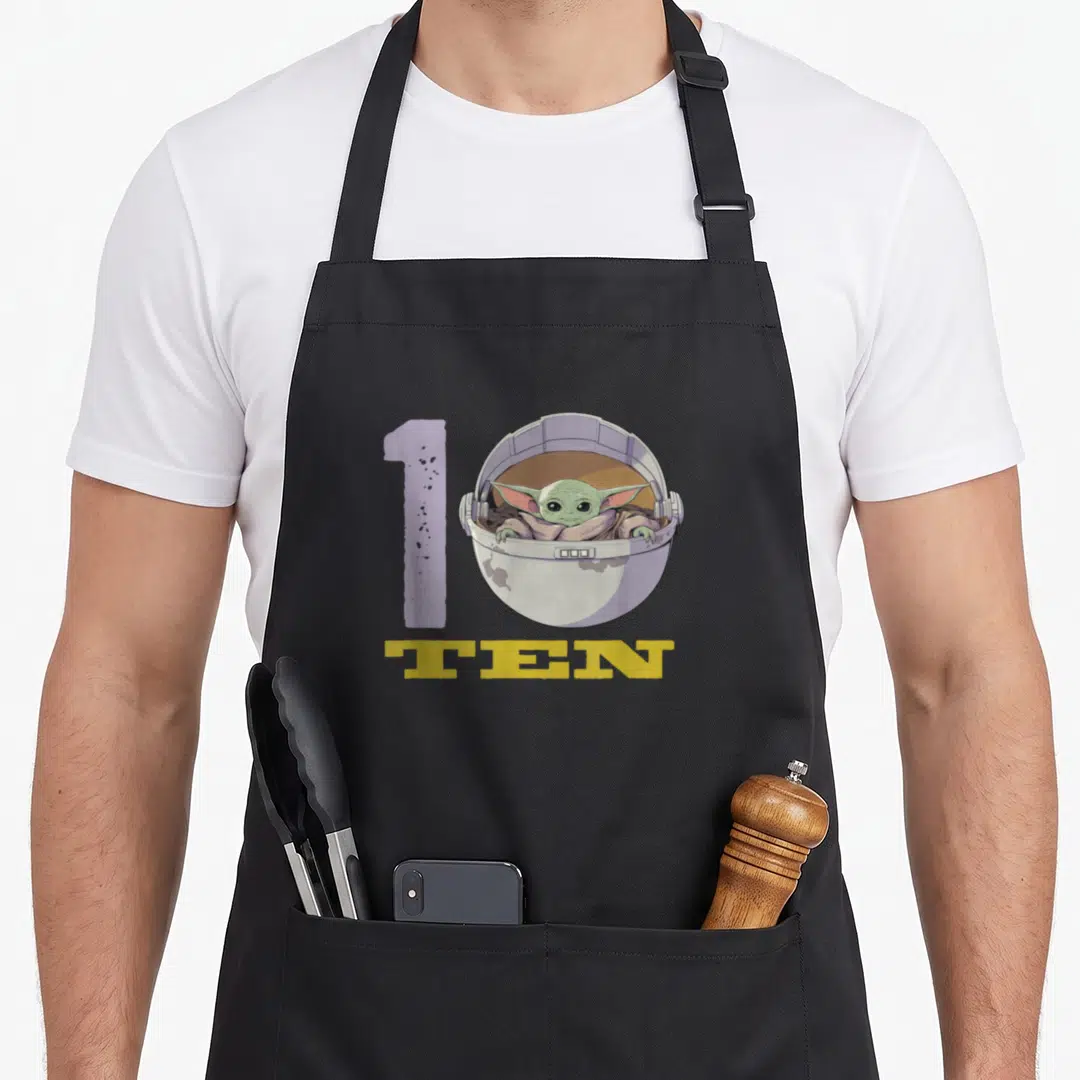 Cute 10th birthday apron - Grogu in floating pod next to purple 1 and yellow TEN text — shown on a black cotton canvas apron against a white background