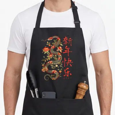Holiday year of the snake apron - Winding creature entwined with flowers and leaves next to vertical Chinese characters — shown on a black cotton canvas apron against a white background