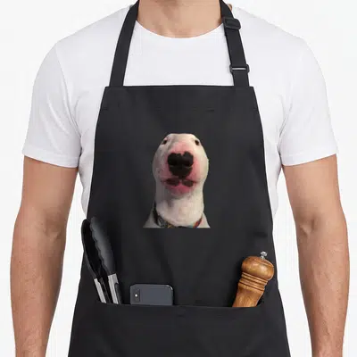 Walter meme apron - White dog face with pink nose and black markings wearing patterned collar — shown on a black cotton canvas apron against a white background