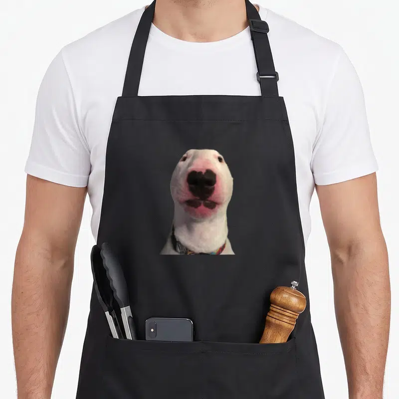 Walter meme apron - White dog face with pink nose and black markings wearing patterned collar — shown on a black cotton canvas apron against a white background