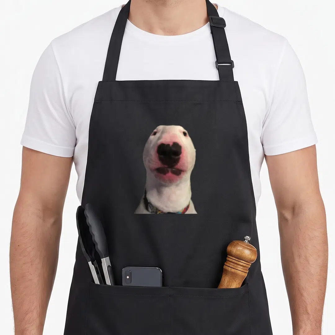 Walter meme apron - White dog face with pink nose and black markings wearing patterned collar — shown on a black cotton canvas apron against a white background