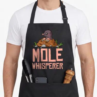 Hunter chemistry apron - Cartoon mole emerging from brown dirt mound with peach text MOLE WHISPERER below — shown on a black cotton canvas apron against a white background