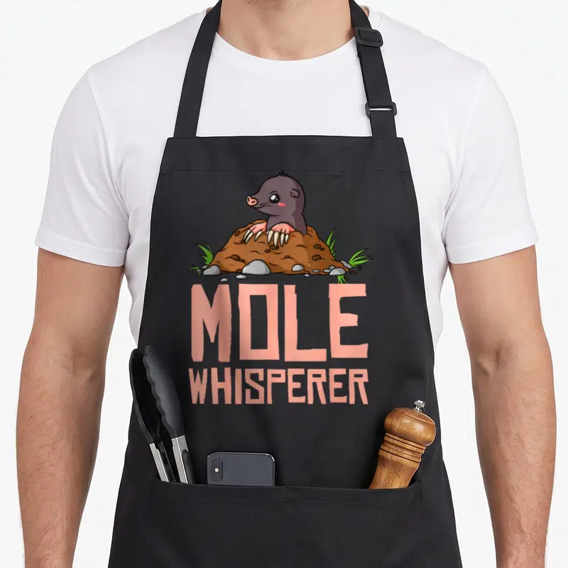 Hunter chemistry apron - Cartoon mole emerging from brown dirt mound with peach text MOLE WHISPERER below — shown on a black cotton canvas apron against a white background