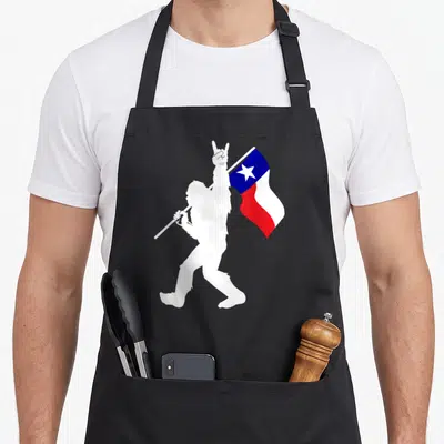 State hunter apron - White bigfoot silhouette walking right holding red white blue Texas flag on pole — shown on a black cotton canvas apron against a white background
