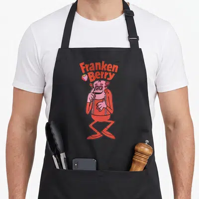 Cereal strawberry apron - Stacked red Franken Berry text above pink cartoon monster with strawberry icon — shown on a black cotton canvas apron against a white background