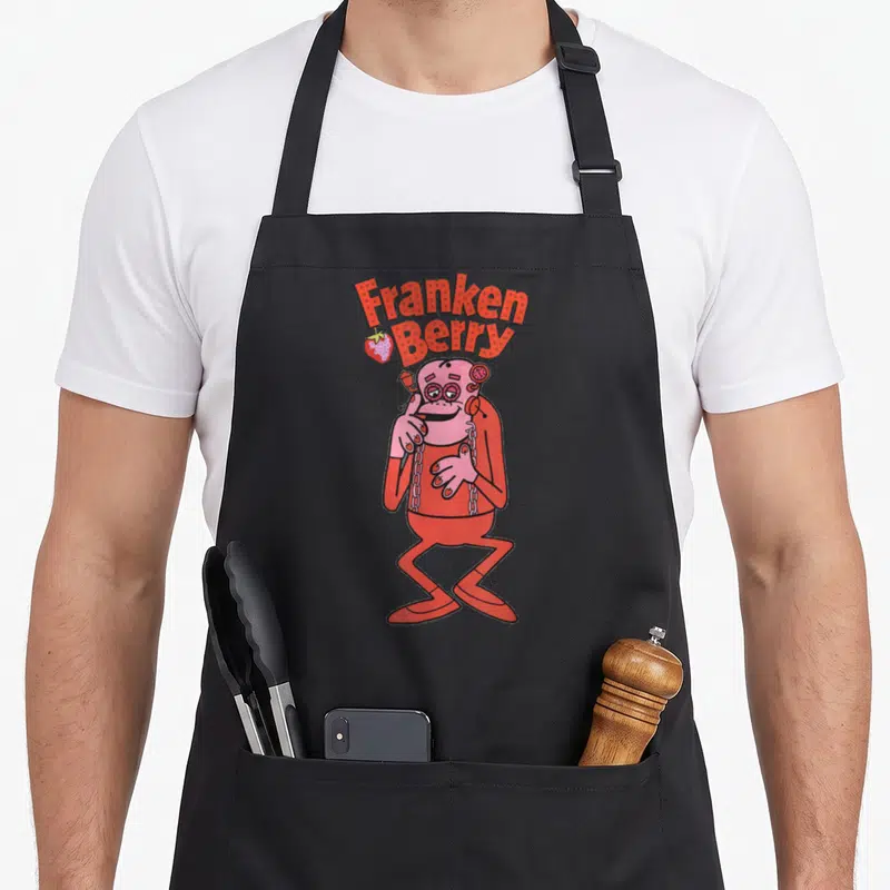 Cereal strawberry apron - Stacked red Franken Berry text above pink cartoon monster with strawberry icon — shown on a black cotton canvas apron against a white background