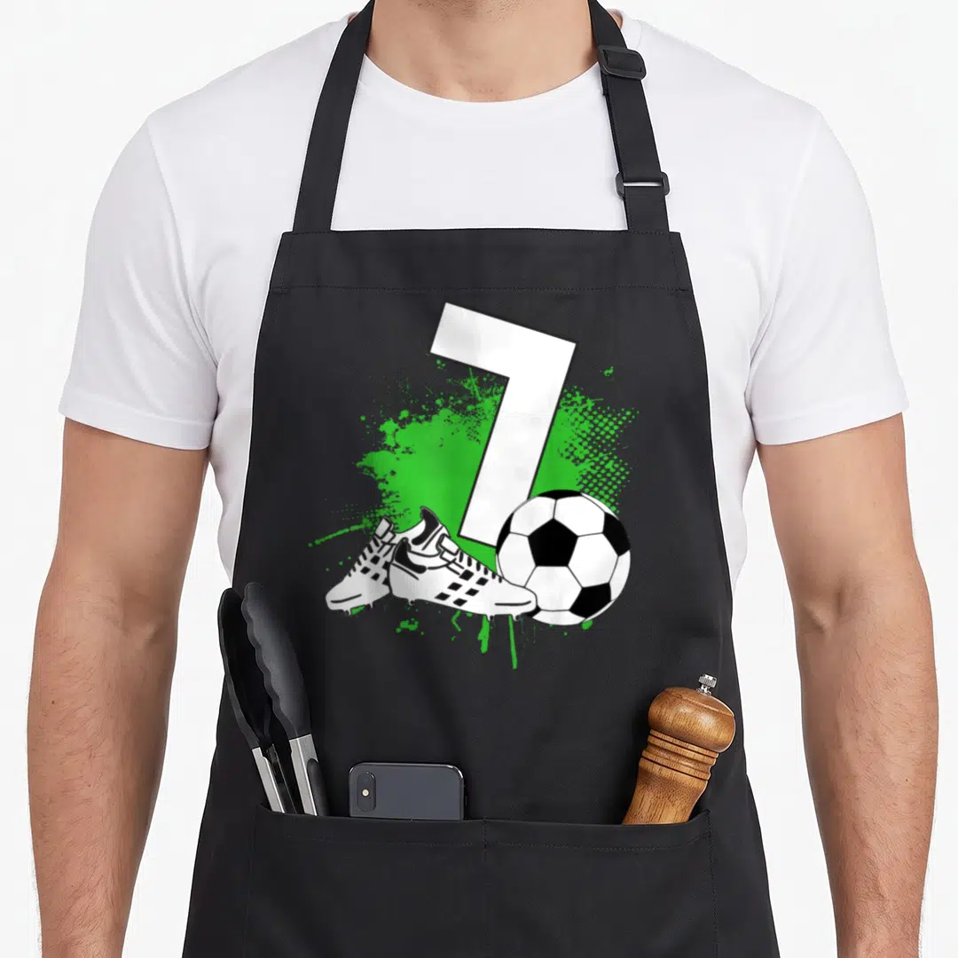Soccer football birthday apron - Number 7 over green splatter with two cleats and soccer ball in cartoon style — shown on a black cotton canvas apron against a white background