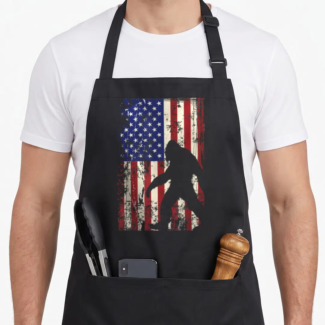 Bigfoot flag 4th of july apron - Grey bigfoot silhouette walking in front of a distressed red white and blue American — shown on a black cotton canvas apron against a white background