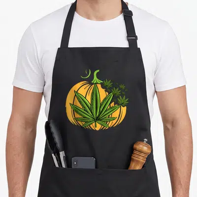Weed veggie apron - Green cannabis leaf over orange pumpkin with smaller leaves in cartoon style — shown on a black cotton canvas apron against a white background