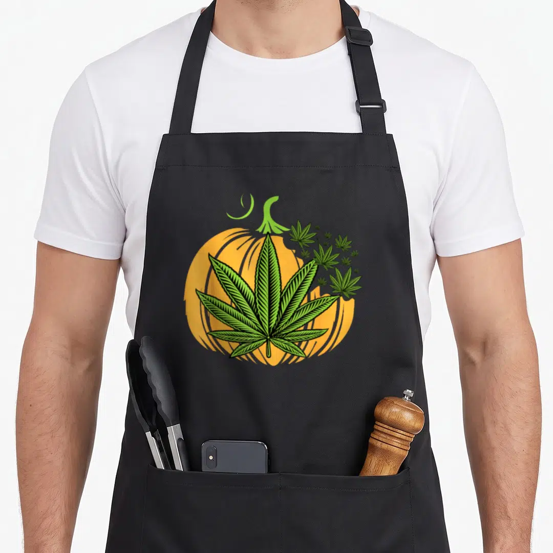 Weed veggie apron - Green cannabis leaf over orange pumpkin with smaller leaves in cartoon style — shown on a black cotton canvas apron against a white background