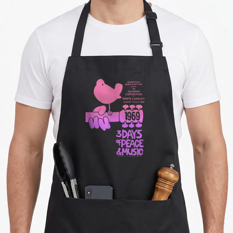 Aquarian woodstock era apron - Pink dove on guitar neck with 1969 Woodstock text and 3 days peace music tagline — shown on a black cotton canvas apron against a white background