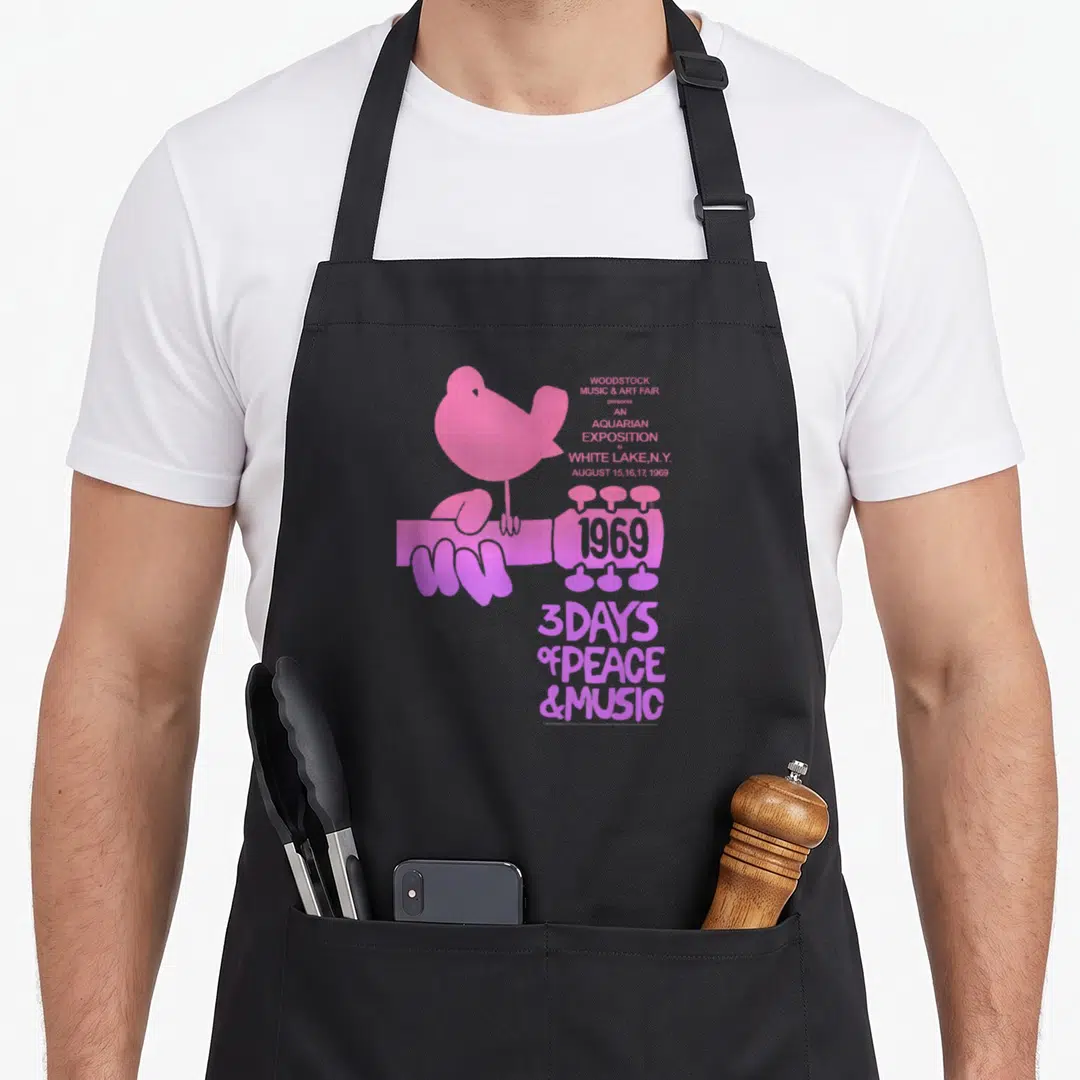 Aquarian woodstock era apron - Pink dove on guitar neck with 1969 Woodstock text and 3 days peace music tagline — shown on a black cotton canvas apron against a white background