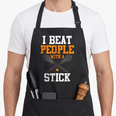 Beat game day apron - I Beat People With A Stick text over crossed lacrosse sticks and ball in white and orange — shown on a black cotton canvas apron against a white background