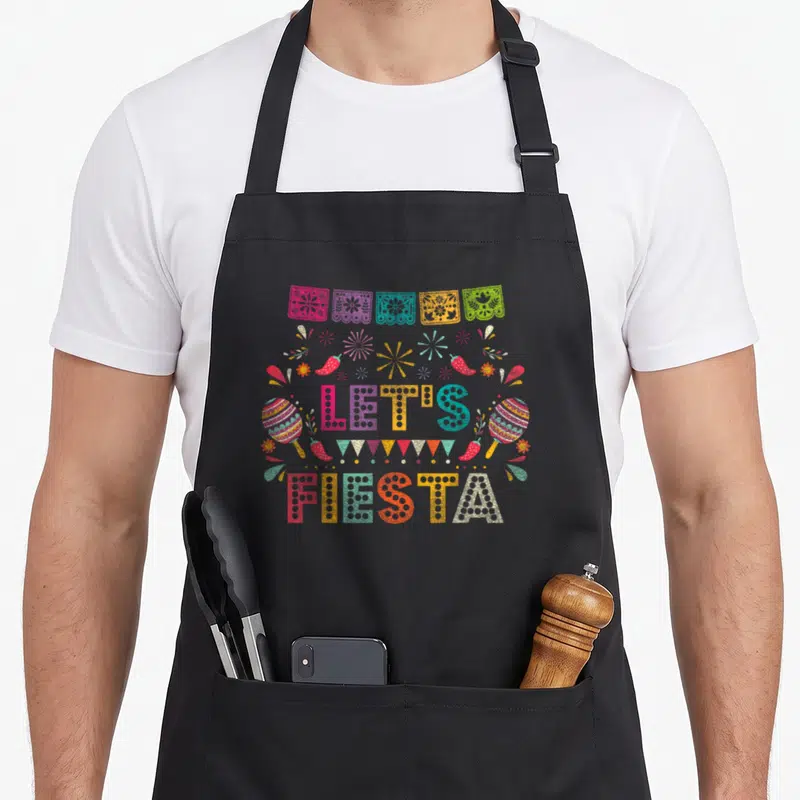 Funny fiesta mexico apron - LET'S FIESTA text with papel picado, maracas, chilies, bunting, and stars in pink blue — shown on a black cotton canvas apron against a white background