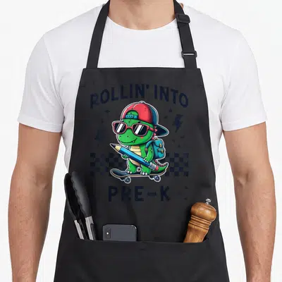 Rollin dinosaur apron - Green dinosaur on skateboard holding pencil with ROLLIN INTO PRE-K text and checkerboard strip — shown on a black cotton canvas apron against a white background