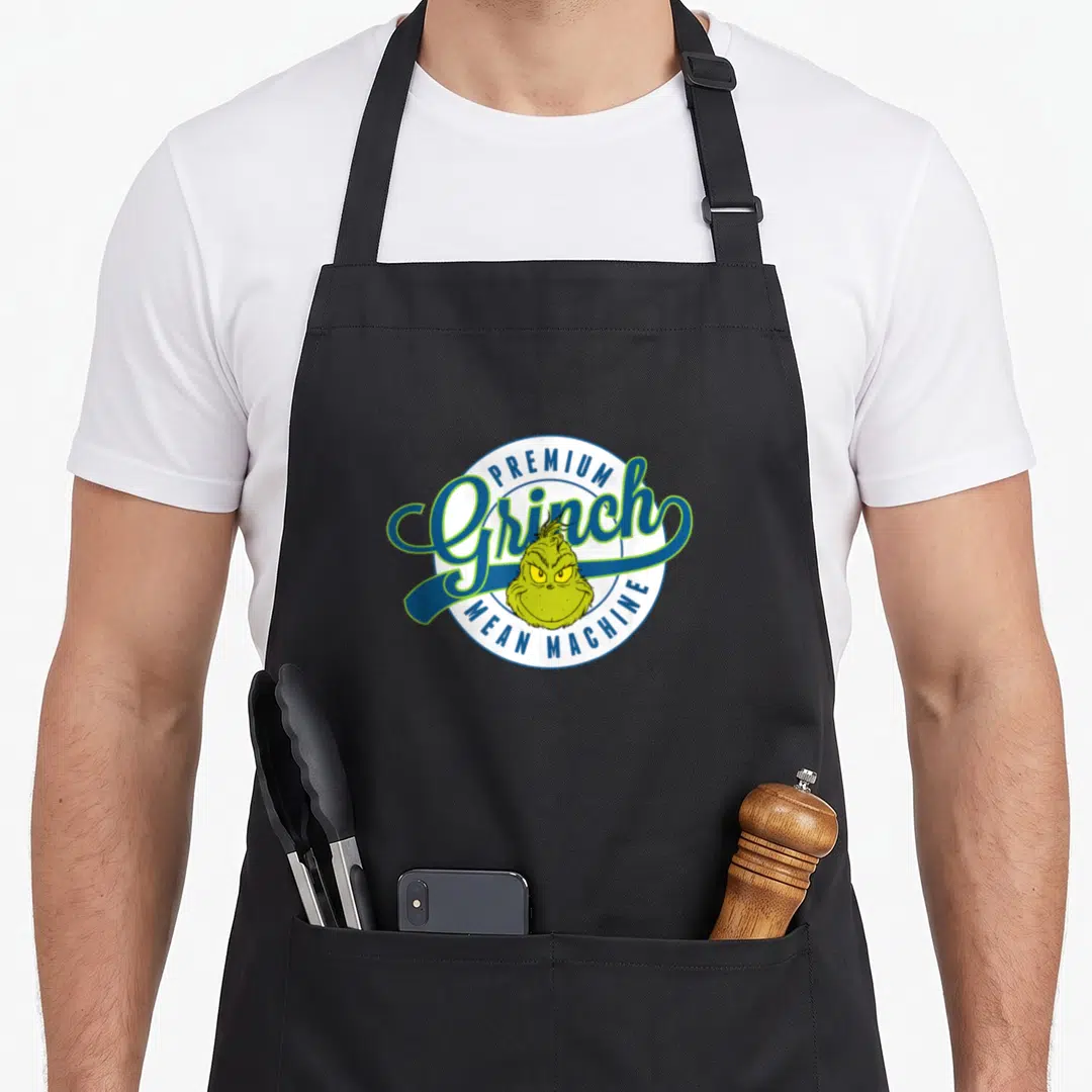 Mean machine apron - Cartoon Grinch head in white circle with blue border and lime green script text — shown on a black cotton canvas apron against a white background