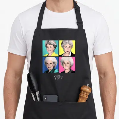Pop golden apron - Four women portraits in pop art grid with cyan, yellow, blue, pink backgrounds and THE GOLDEN GIRLS — shown on a black cotton canvas apron against a white background