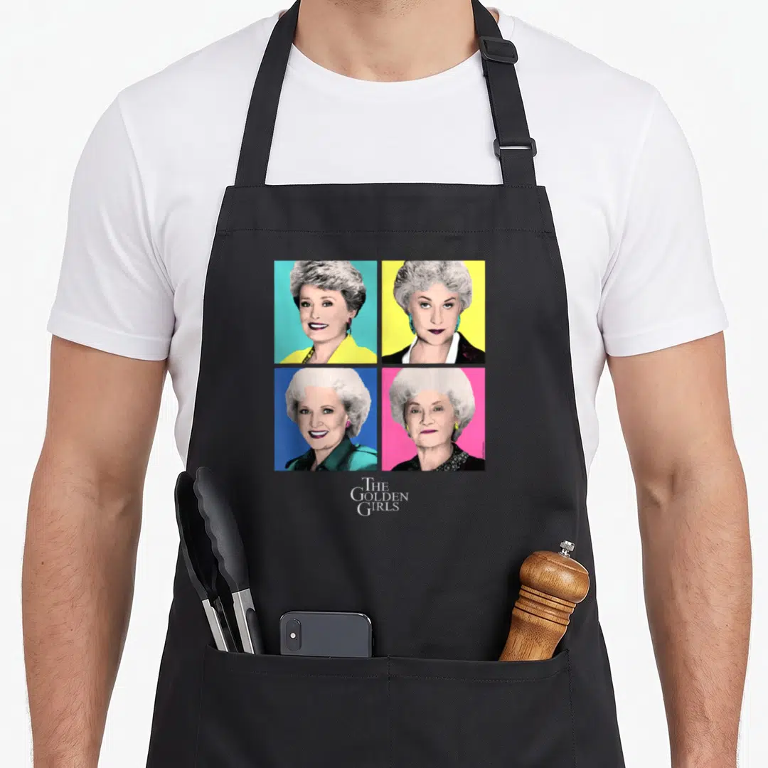 Pop golden apron - Four women portraits in pop art grid with cyan, yellow, blue, pink backgrounds and THE GOLDEN GIRLS — shown on a black cotton canvas apron against a white background