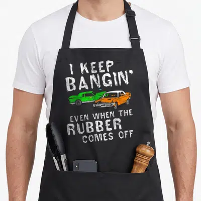 Bangin rubber apron - Two crashed cartoon cars in green and orange with white text about demolition derby — shown on a black cotton canvas apron against a white background