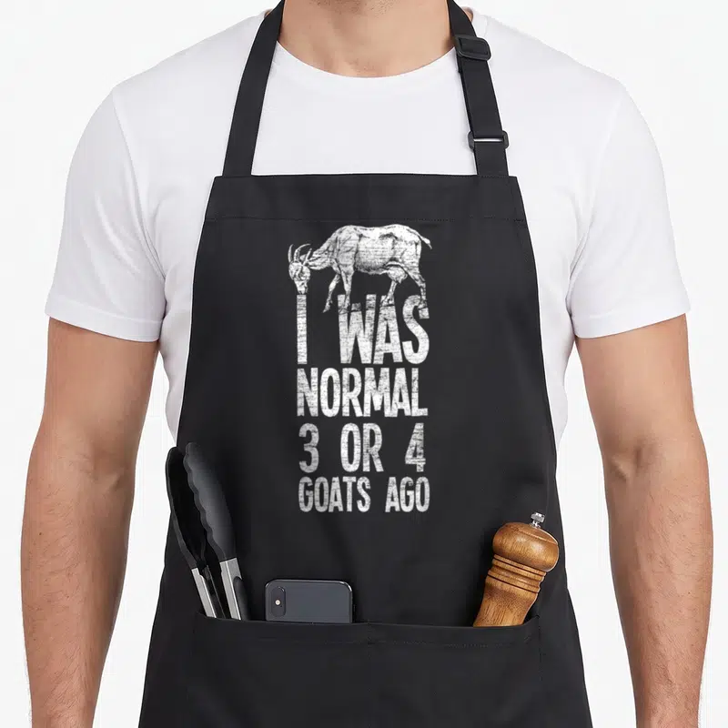 Three goat apron - White distressed goat illustration above stacked text I WAS NORMAL 3 OR 4 GOATS AGO — shown on a black cotton canvas apron against a white background