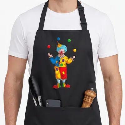 Juggling clown aprons - Cartoon clown juggling four red, yellow, blue, and green balls in a circus pose — shown on a black cotton canvas apron against a white background