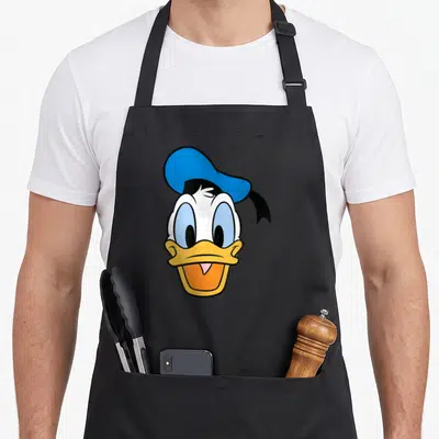 Caballero face apron - Donald Duck head with blue hat, yellow beak, orange mouth, white face, cartoon style — shown on a black cotton canvas apron against a white background