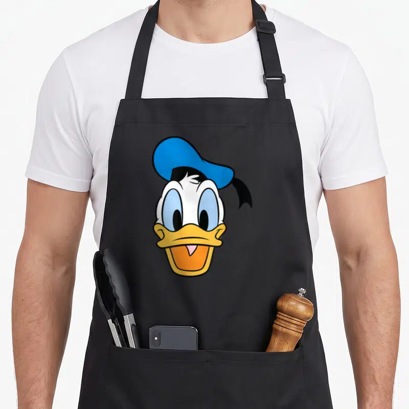 Caballero face apron - Donald Duck head with blue hat, yellow beak, orange mouth, white face, cartoon style — shown on a black cotton canvas apron against a white background
