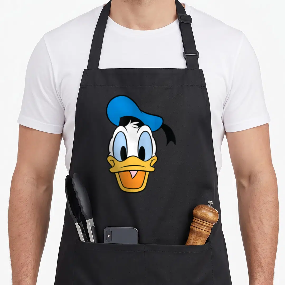 Caballero face apron - Donald Duck head with blue hat, yellow beak, orange mouth, white face, cartoon style — shown on a black cotton canvas apron against a white background